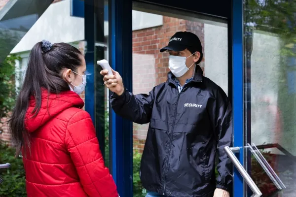 An unarmed security guard checks visitors’ body temperature at an entrance.